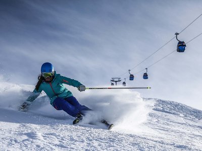 Ausflugsziele in Sölden gesucht? Skifahrerin in blauer Ausrüstung fährt auf frischem Schnee mit Seilbahn im Hintergrund