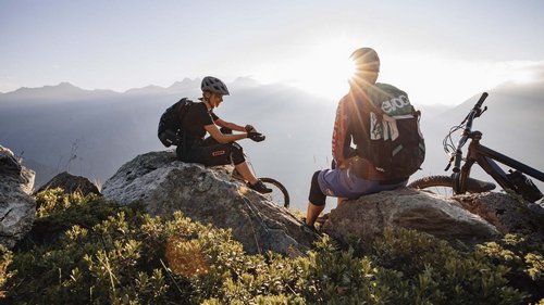 Ausflugsziele in Sölden gesucht? Zwei Mountainbiker machen Pause auf Felsen mit Blick auf Berge und Sonnenuntergang.