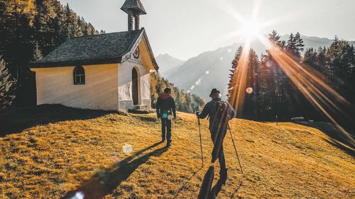 Ausflugsziele in Sölden gesucht? Zwei Wanderer gehen bei Sonnenuntergang an einer kleinen Kapelle auf einem Hügel vorbei