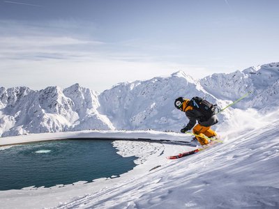 Ausflugsziele in Sölden gesucht? Skifahrer fährt einen schneebedeckten Hang neben einem runden, teilweise zugefrorenen Wasserbecken in den Bergen