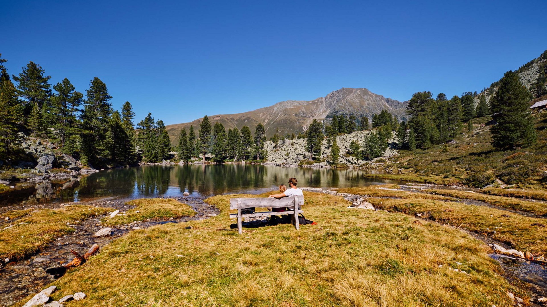 Dein 4-Sterne-Hotel im Ötztal Personen sitzen auf Bank mit Blick auf Bergsee und Wälder im Sonnenschein