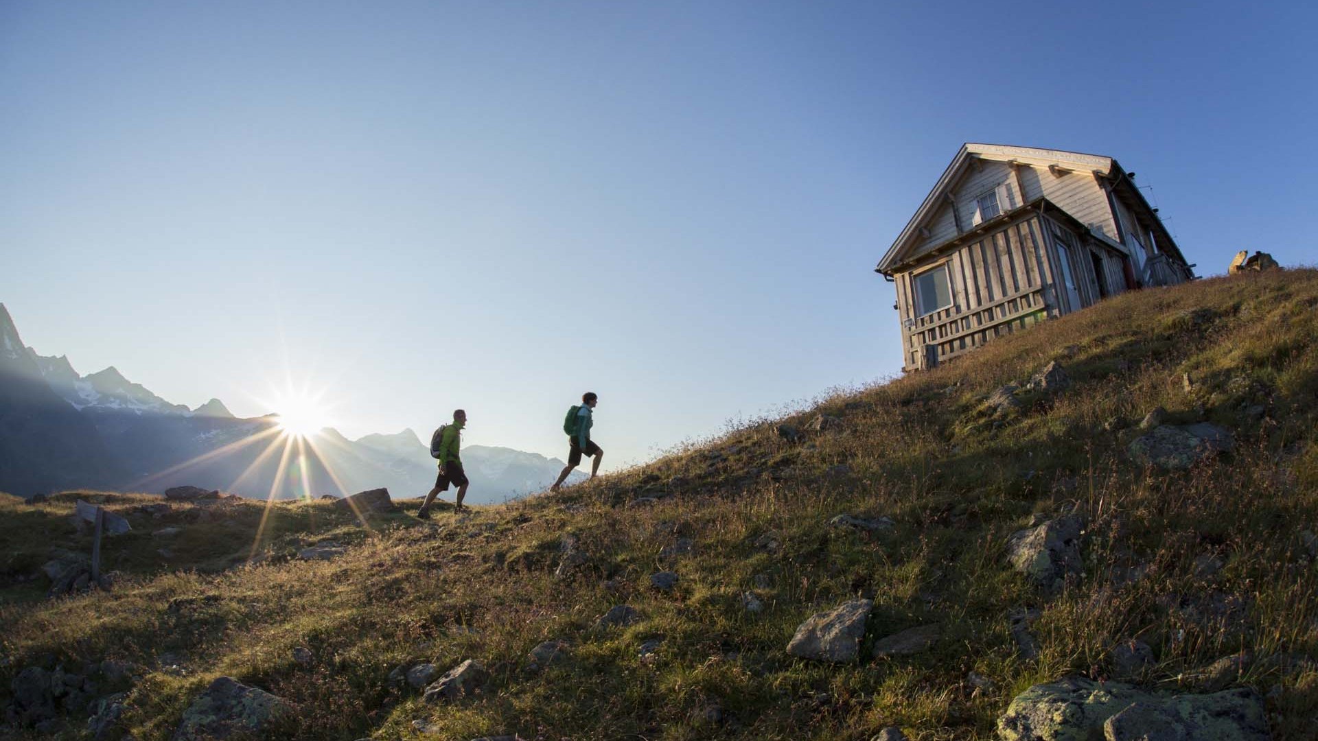 Wandern in Sölden? Im Sommer und Winter! Zwei Wanderer gehen bei Sonnenaufgang zu einer Berghütte auf einem Hügel