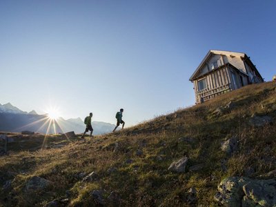 Wandern in Sölden? Im Sommer und Winter! Zwei Wanderer gehen bei Sonnenaufgang zu einer Berghütte auf einem Hügel